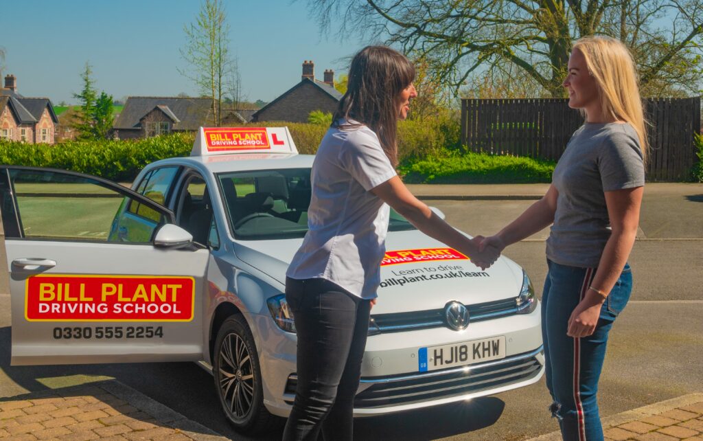 A female driving instructor shaking hands with a student in front of a car.